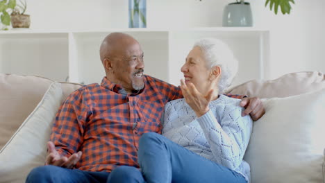 portrait of happy senior biracial couple sitting on couch embracing and talking at home, slow motion