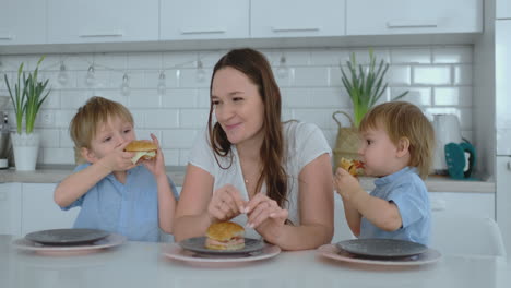 A-young-beautiful-mother-in-a-white-dress-with-two-children-is-smiling-and-eating-fresh-burgers-in-her-kitchen.-Happy-family-homemade-food-healthy-foods