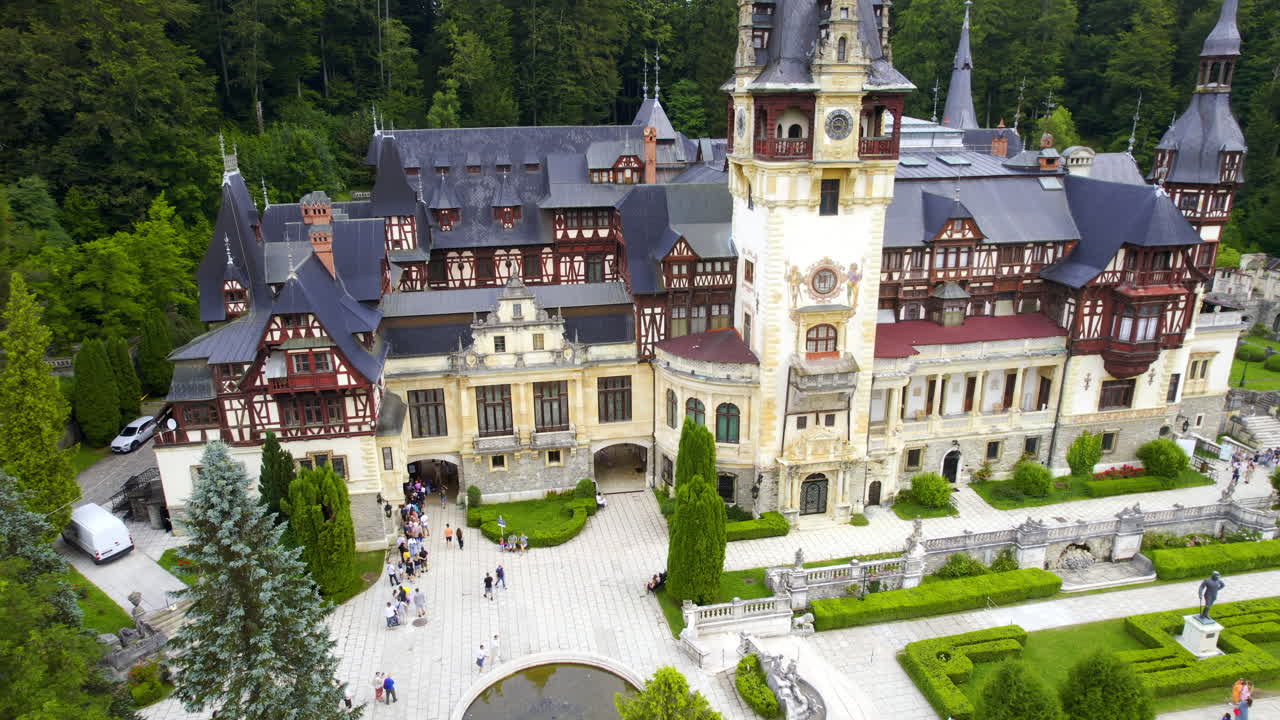Luftfoto med drone af Peleș-slottet i Rumænien. Slot med haver og turister i Karpaterne, frodig skov omkring.