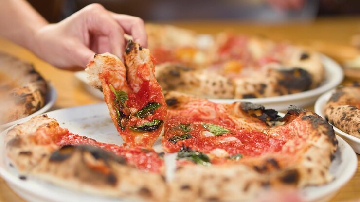 Primo piano di una mano che solleva una fetta di pizza a crosta sottile con salsa di pomodoro e basilico.