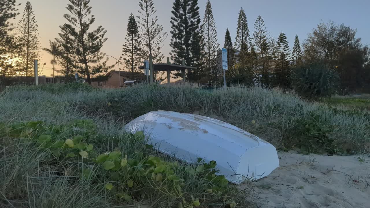 A white boat lay upside down moored on the bank of a beach with a sunset background.