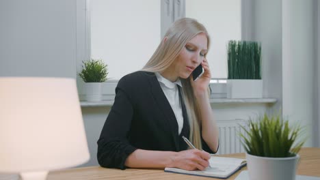 business woman talking on smartphone in office. elegant young blond female in office suit sitting at workplace and negotiating via mobile phone in hand writing down necessary information into notebook