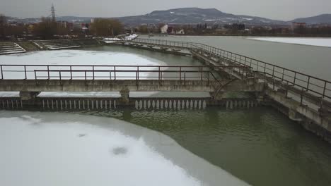 Frozen-lake-in-winter,-aerial-drone-shot-from-above,-concrete-walkways-in-lake,-dark,-cloudy-and-moody-scene,-revealing-shot,-town-in-background,-revealing-shot