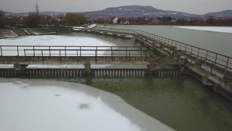 lago congelado en invierno, toma aérea de drones desde arriba, pasarelas de concreto en el lago, escena oscura, nublada y malhumorada, toma reveladora, ciudad en segundo plano, toma reveladora