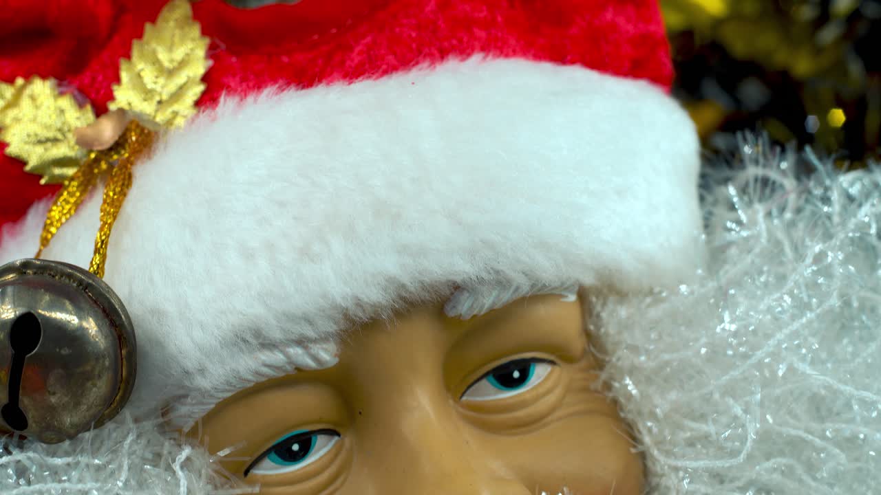 Hračka Santa Clause v červené čepici se zvonečky, velký bílý vous, vánoční dekorace, tradiční sváteční dárky, novoroční výzdoba, lesklé barevné uspořádání, kreativní detailní plynulý záběr s náklonem dolů, 4K video.