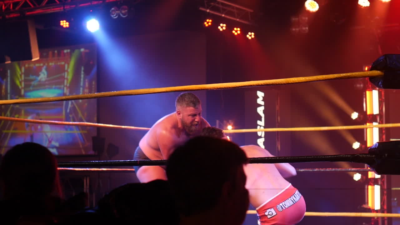 A wrestler hoists his opponent over his shoulder in a dramatic lift inside the wrestling ring, with the opponent’s legs kicking mid-air.