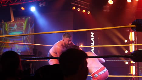 a wrestler hoists his opponent over his shoulder in a dramatic lift inside the wrestling ring, with the opponent’s legs kicking mid-air.