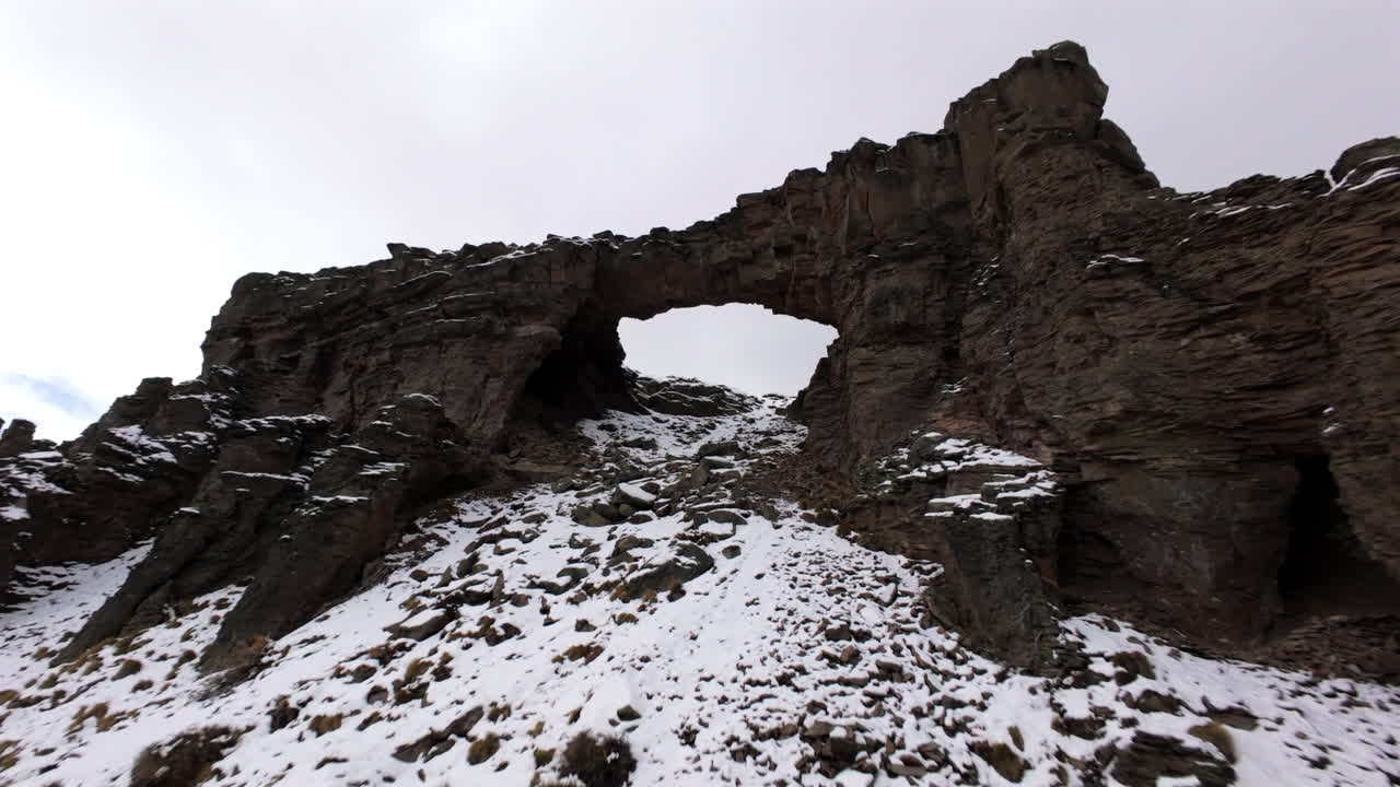 Viaje inmersivo FPV rozando las aguas del Lago Caviahue, pasando columnas de basalto cubiertas de nieve y elevándose a través de un famoso puente de piedra volcánico y accidentado en los Andes, capturado en movimiento aéreo dinámico.