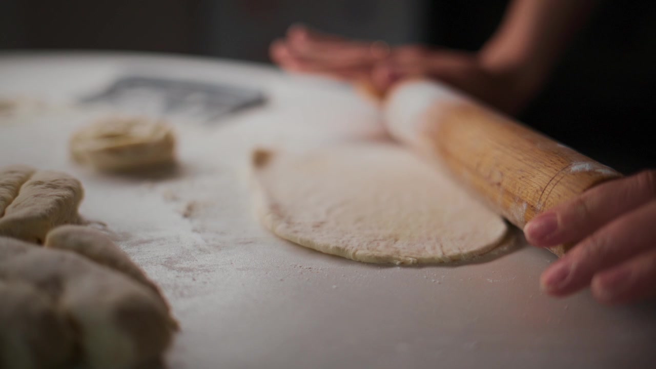 Mani stendono l'impasto su una superficie infarinata. Le dita guidano con cura un mattarello di legno. L'impasto si assottiglia mentre la farina si sparge nell'aria. L'attenzione rimane sul movimento abile, non sul viso. Perfetto per tutorial di cucina.