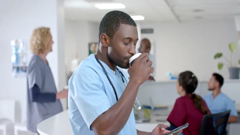 Portrait-of-african-american-male-doctor-drinking-coffee,-using-smartphone-in-corridor,-slow-motion