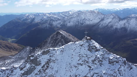 Aerial-arc-shot-of-a-cross-at-the-summit-of-the-Iceman's-Otzi-Peak,-a-breathtaking-panoramic-viewing-point-surrounded-by-the-picturesque-snow-capped-mountains-of-the-Val-Senales-Glacier,-Tyrol,-Italy