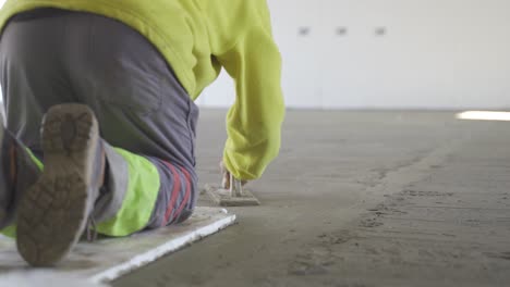 Vista-Estática-De-Un-Trabajador-Que-Nivela-El-Piso-Enyesado-Usando-Paleta-Plana-Y-Cemento-Manualmente-Usando-Una-Chaqueta-De-Neón-Y-Pantalones-Grises