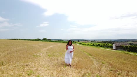 young pregnant woman in a white summer dress walking down a field holding her belly and flowers for a maternity photo shoot