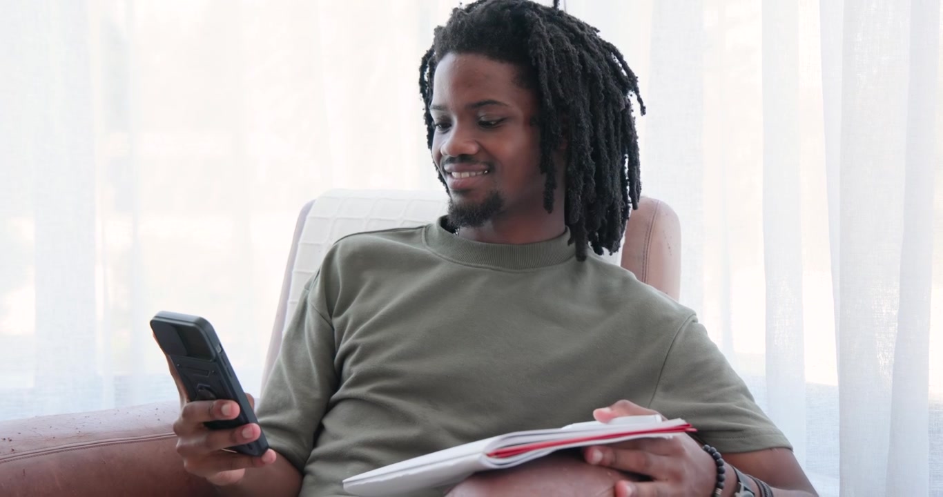 African American man writing on notebook with pen on couch in green shirt, pausing, checking phone
