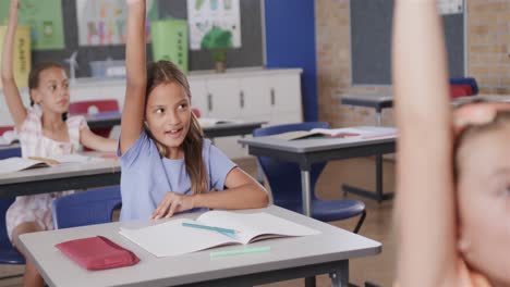 Colegialas-Felices-Y-Diversas-Levantando-La-Mano-En-Los-Escritorios-De-La-Clase-De-Escuela-Primaria,-Cámara-Lenta