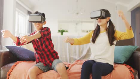 happy asian brother and sister at home, sitting on couch in living room wearing vr headsets