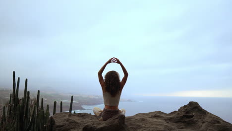 With-the-ocean-and-green-mountains-in-view,-a-woman-meditates-on-a-mountain's-peak,-accompanied-by-the-Maha-Sakal-hand-gesture