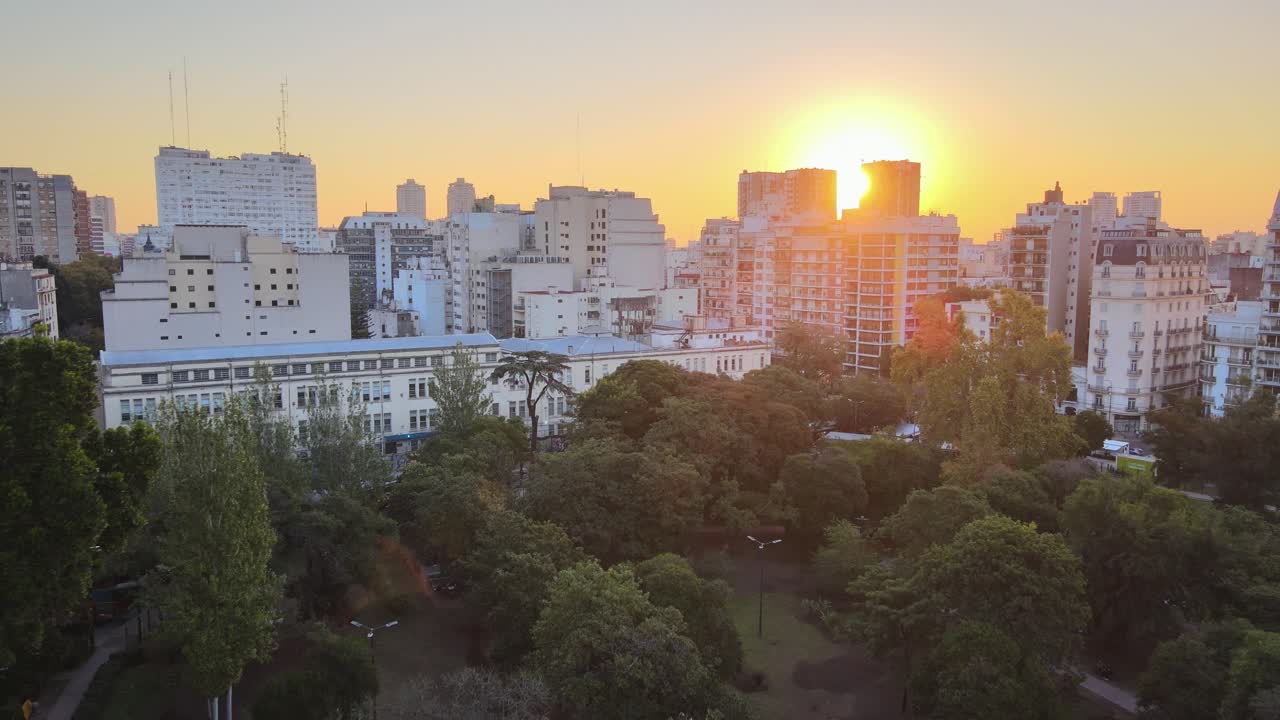 Sun setting behind mid rise buildings in Buenos Aires with green treetops of Parque Rivadavia in foreground. Argentina, dolly in aerial
