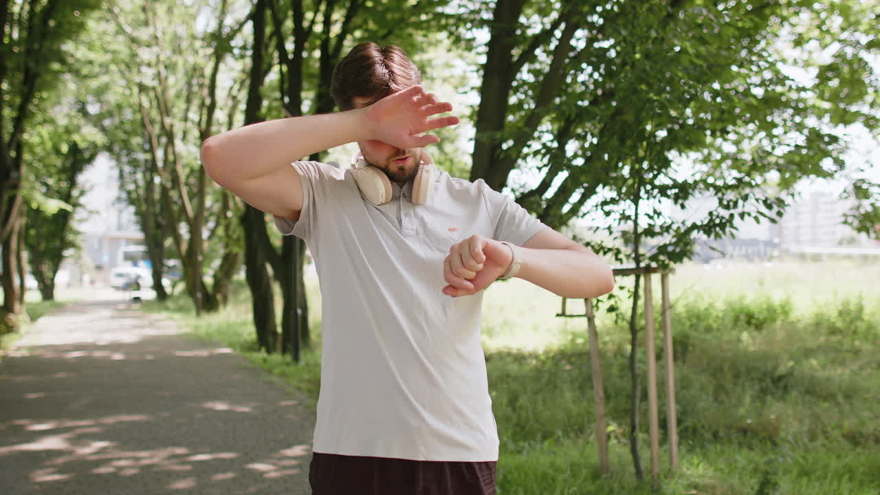 Active young man jogger running in public park and looking at smartwatch getting tired relaxing