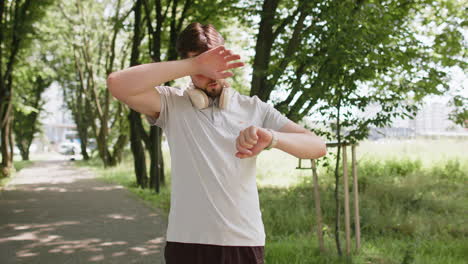 active young man jogger running in public park and looking at smartwatch getting tired relaxing
