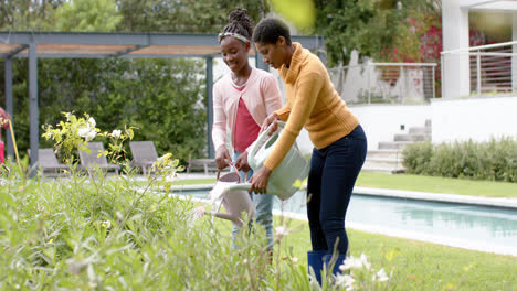 happy african american daughter and mother watering plants in sunny garden, slow motion