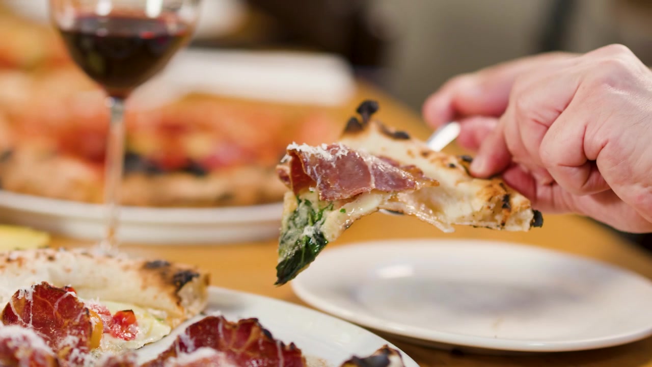 Primo piano di mani che sollevano una fetta di pizza al prosciutto su un piatto accanto a del vino rosso.