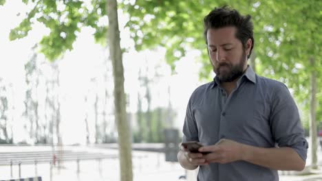 Hombre-Barbudo-Enfocado-Usando-Un-Teléfono-Inteligente-Al-Aire-Libre