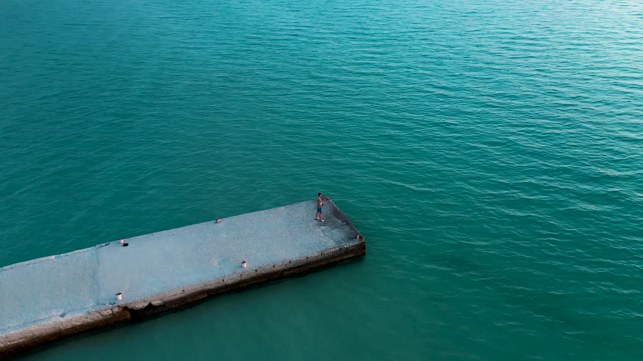 Einführende Kreisaufnahme eines Mannes auf einem Steg, der auf die Sonne und das klare blaue Meer blickt.