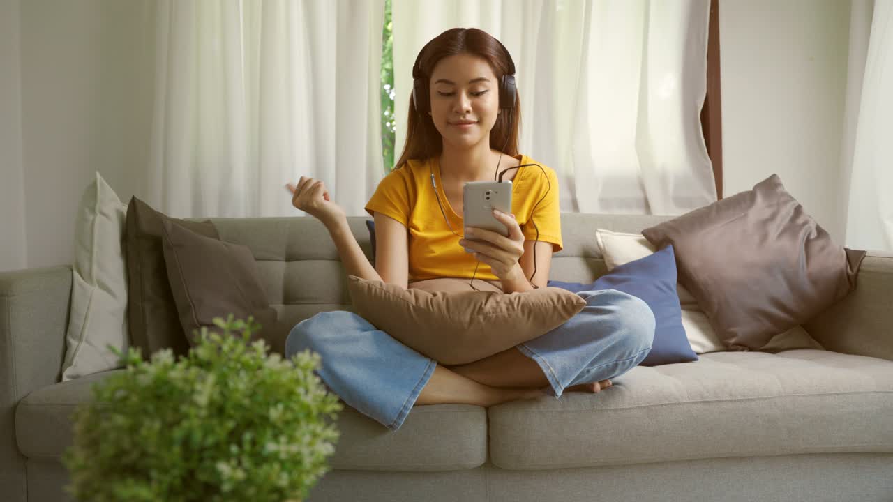 An woman Smiling is listening to music through headphones and using smartphone. Lady relaxing on comfortable sofa with eyes closed at home. asian teen Happy and feeling no stress.