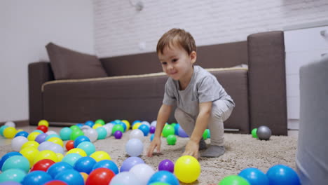 kid in a grey t-shirt sits on the floor in his room. lovely toddler jumps among the balls scattered by the carpet.