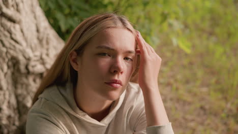 young girl seated on ground resting her head on left hand, gently touching her hair with a thoughtful, contemplative expression, background features tree bark, foliage, and greenery