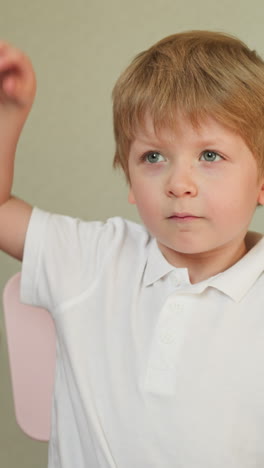 calm blond boy sits at desk listening attentively. preschooler raises hand and exclaims answering teacher question at kindergarten lesson slow motion