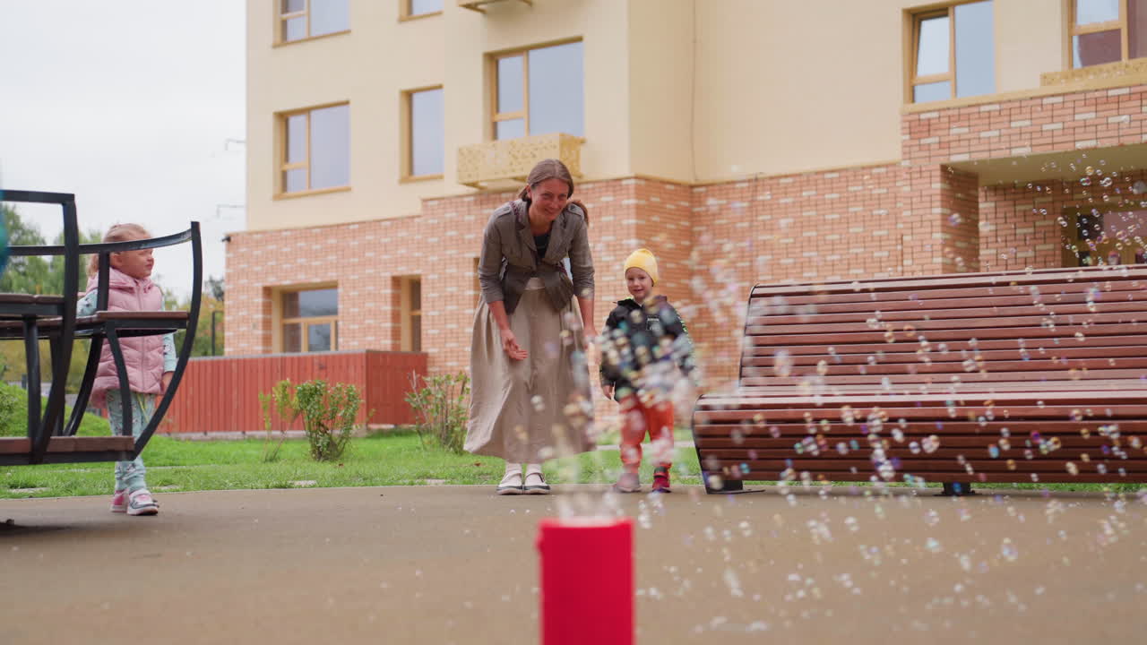 Smiling mother and children standing outdoors in courtyard watching colorful bubbles float from soap machine, showing family excitement, playful curiosity, and joyful bonding