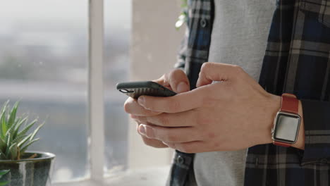 Cerrar-Las-Manos-De-Una-Mujer-Usando-Un-Teléfono-Inteligente-En-Casa-Enviando-Mensajes-De-Texto-Escribiendo-Mensajes-De-Correo-Electrónico-Navegando-En-Las-Redes-Sociales-En-Línea-De-Pie-Junto-A-La-Ventana