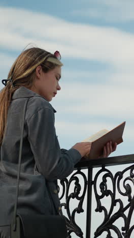 hermosa mujer con ropa gris descansa su mano en una valla de hierro mientras hojea un libro en su mano, el viento sopla suavemente su cabello, y su ropa aleta suavemente