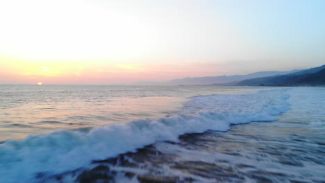 Aerial-shot-of-an-ocean-wave-breaking-on-the-beach-under-a-soft-sunset-sky-background-down-the-coast-of-Ventura,-California-DRONE