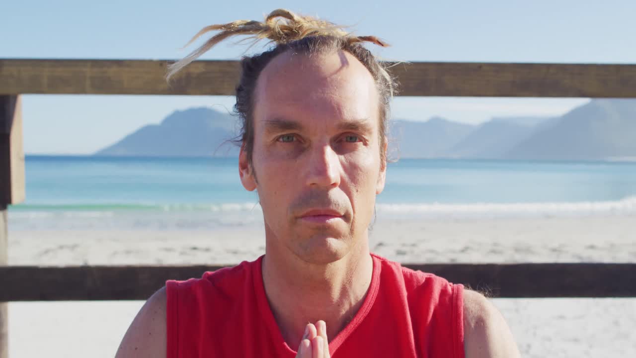 retrato de video de un hombre caucásico tranquilo con rastas practicando meditación de yoga en la playa al sol