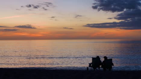 Pareja-Sentada-En-Sillas-En-La-Playa-Viendo-La-Puesta-De-Sol