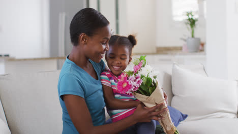 Happy-african-american-mother-and-daughter-sitting-on-sofa-and-smelling-flowers