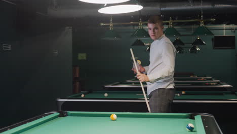 man in white shirt and grey trousers holds cue stick and glass of lemon drink while pacing around billiards room. overhead lights cast a moody ambiance, enhancing the stylish and relaxed atmosphere