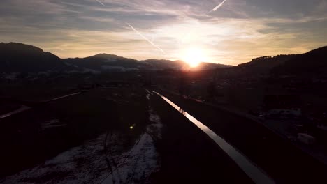 Beautiful-moody-winter-wonderland-scenery-in-Switzerland-with-a-reflective-river,-cars-and-a-village-while-sunset