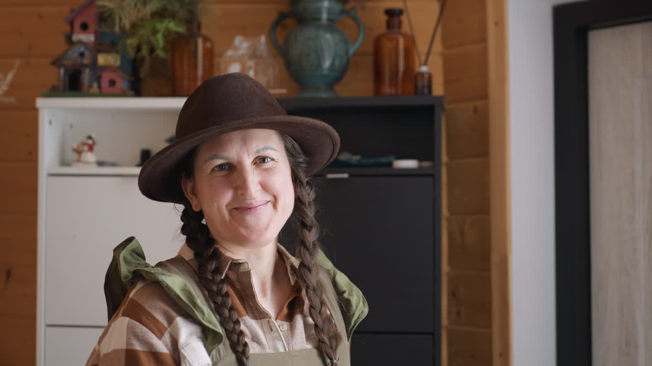 Portrait of cheerful lady smiling behind cupboard topped with flower and candle vases in wooden room wearing olive gown, plaid shirt, brown hat under warm natural light, with braided hair