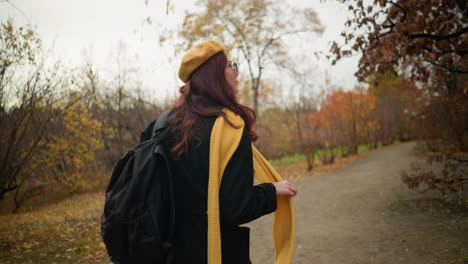 back view of woman wearing yellow muffler and beret walking through forest, holding muffler while looking to side, with hair swaying gently in autumn breeze