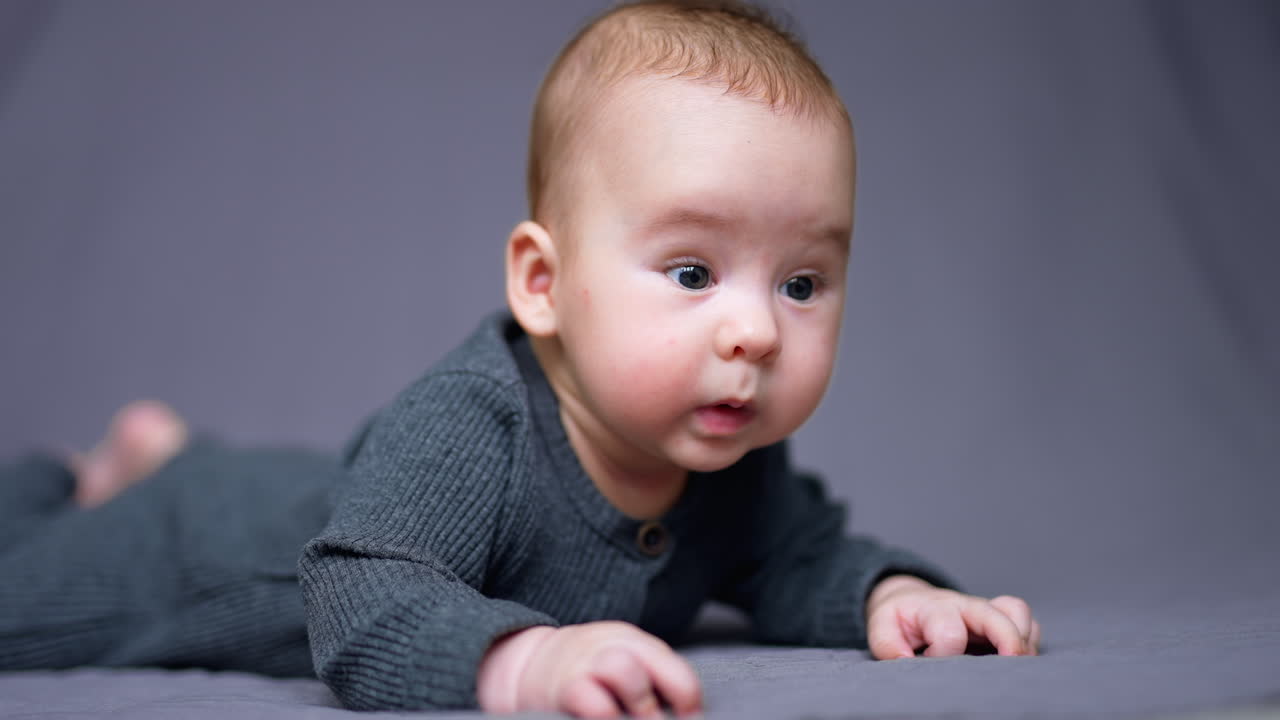 Little cute baby boy in grey outfit rests on his belly. Lovely child looks sideways with interest. Grey blurred backdrop.