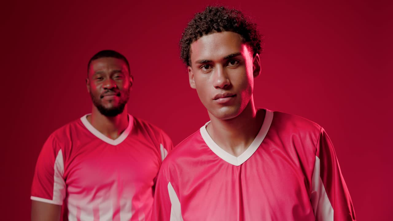 Primer plano de hombres afroamericanos con uniforme sosteniendo un balón de fútbol en un set rojo, la cámara se acerca mientras avanzan.