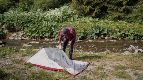 una revisión de un tipo con una camisa a cuadros roja que monta una tienda de campaña en su caminata cerca de un río de montaña en el bosque
