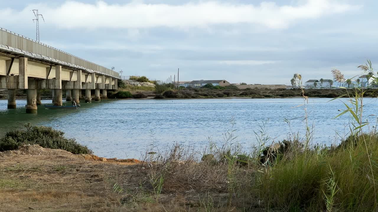 Static view of a fisherman in a motorboat passing under a bridge. Daylight