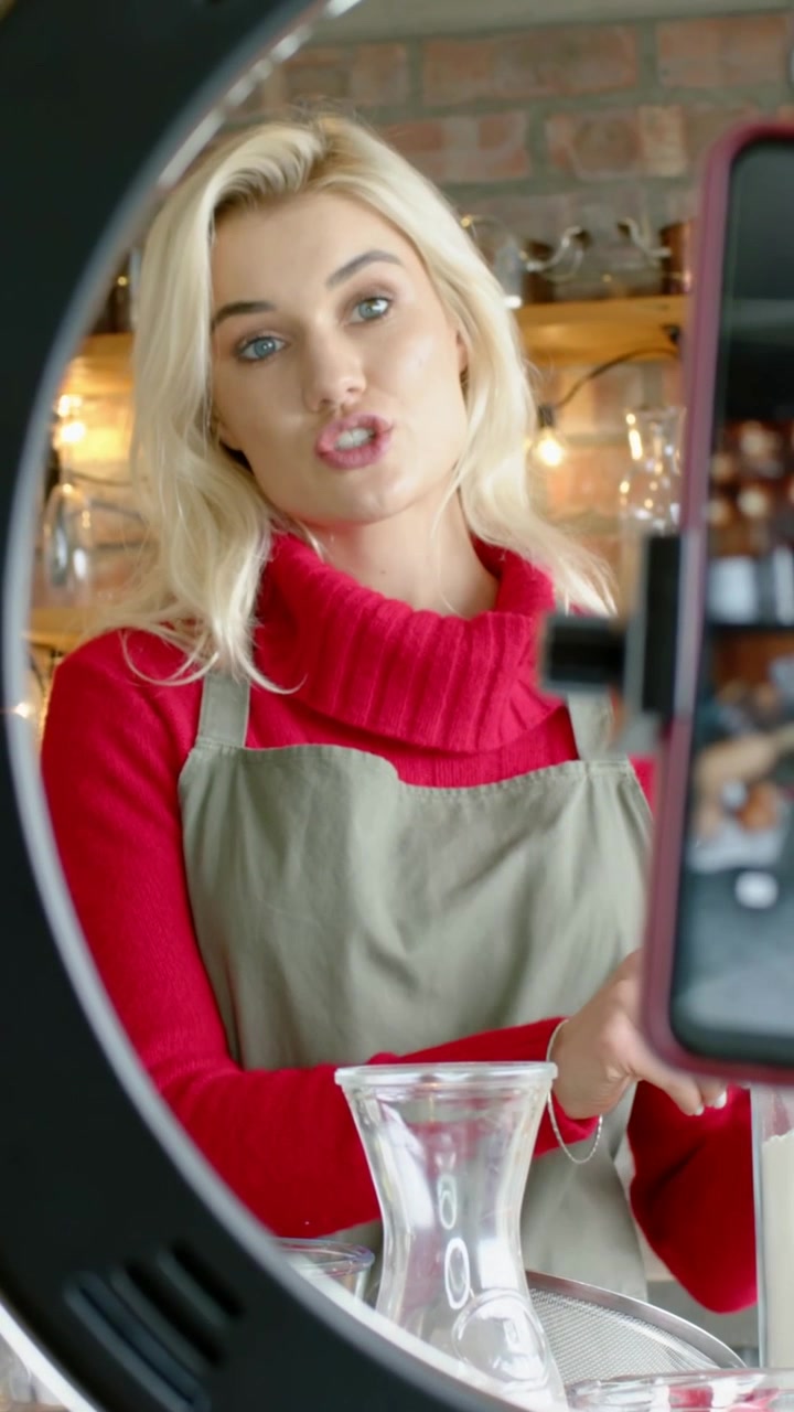 Video verticale: Telefono che registra una donna con maglietta rossa e grembiule verde oliva che versa latte in una caraffa per una dimostrazione in caffetteria.