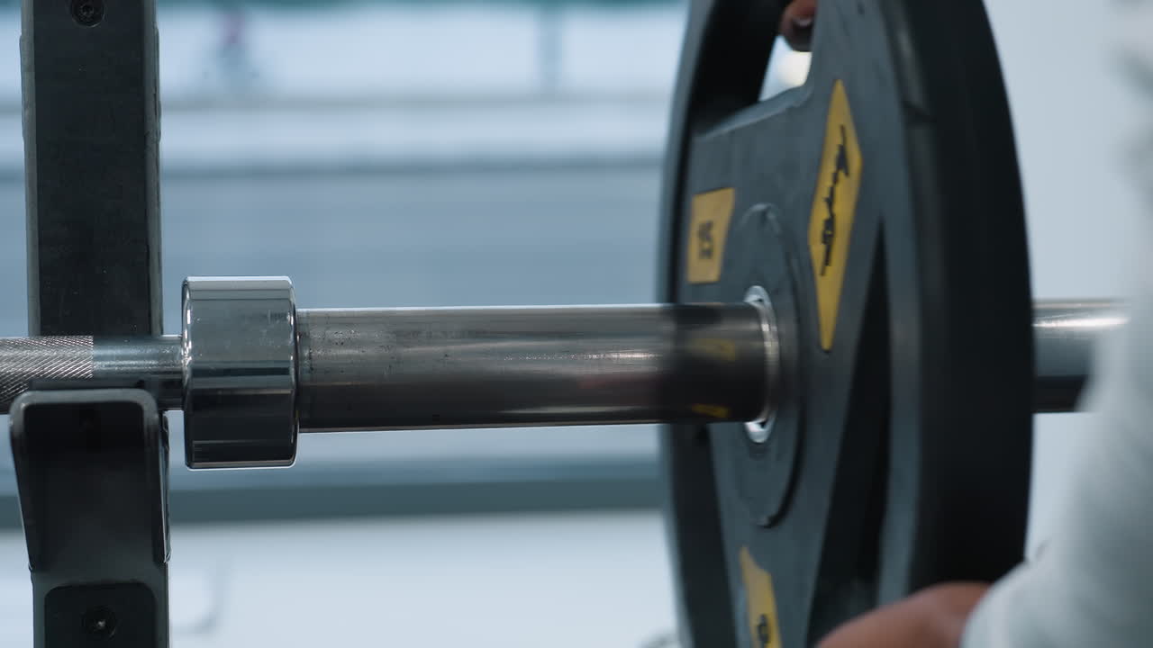 Close up of person clipping metal weight plate onto barbell sleeve beside window with blurred car passing by, showing hands securing heavy plate on chrome bar during gym equipment