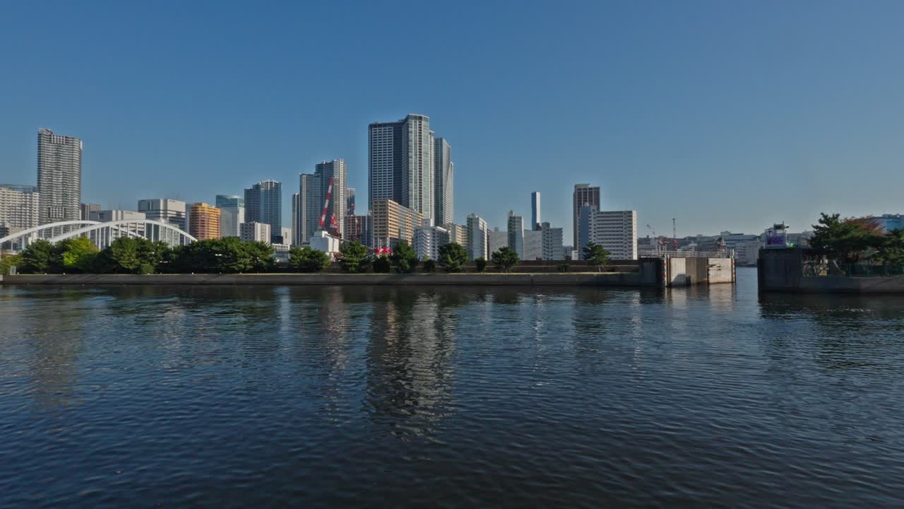 A panoramic view of Tokyo's modern skyline, featuring numerous high-rise buildings and a bridge, seen across a calm body of water.